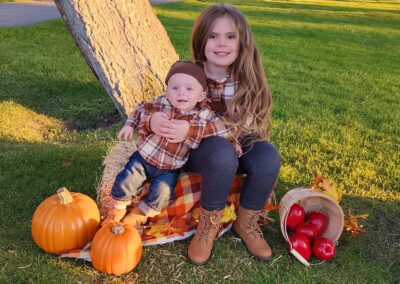 Male infant snuggling with older sister on a straw bale with pumpkins and a bushel of apples on the ground beside them